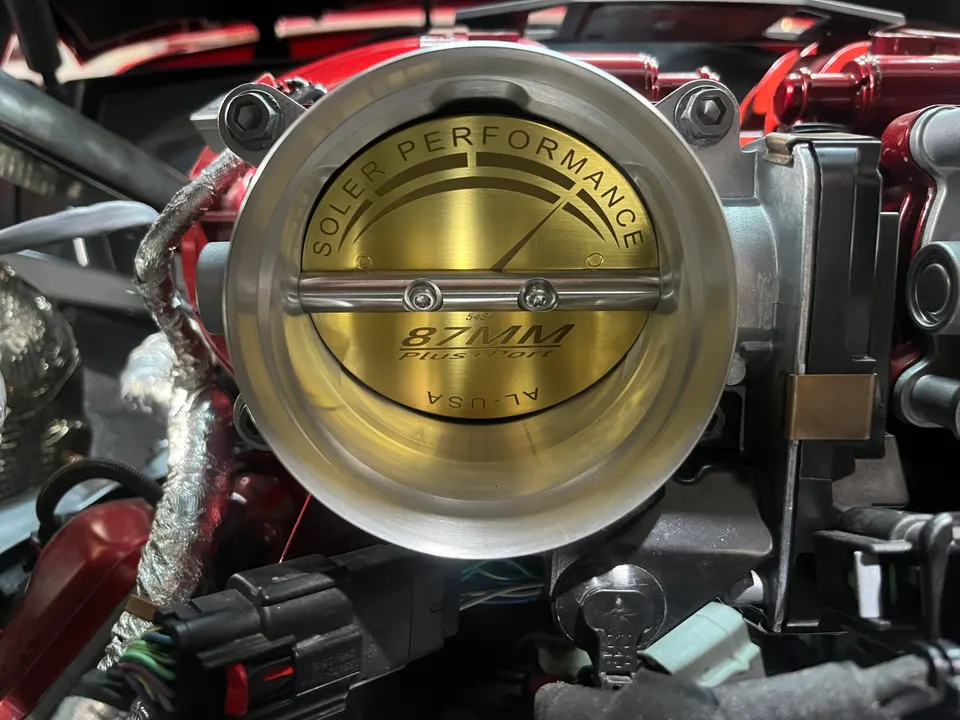 Close-up of a Soler Performance 87mm throttle body installed in a car's engine bay, showcasing the gold-colored valve.