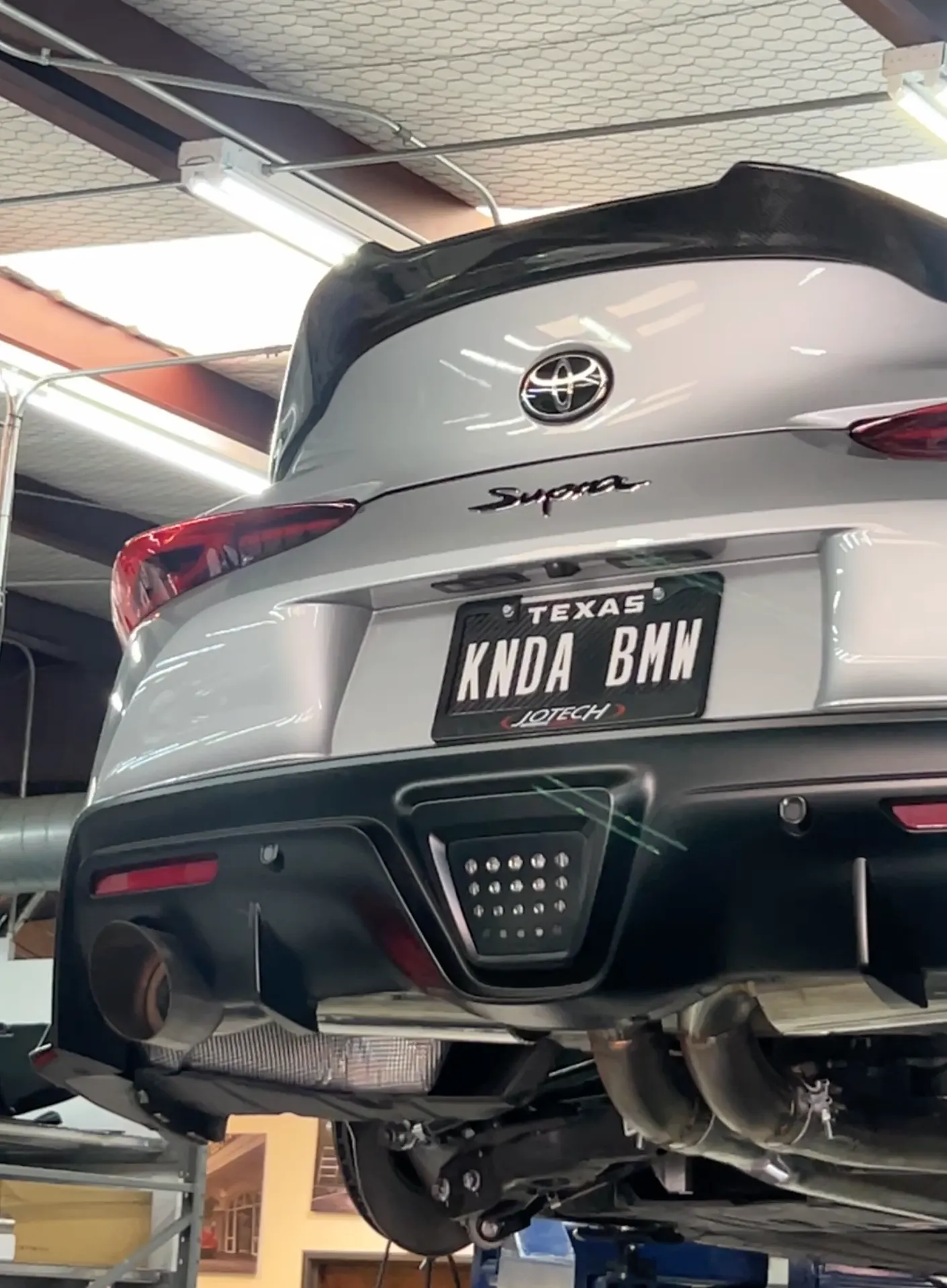 Rear view of a silver Toyota Supra on a lift in a shop, showcasing its aftermarket exhaust system and diffuser.