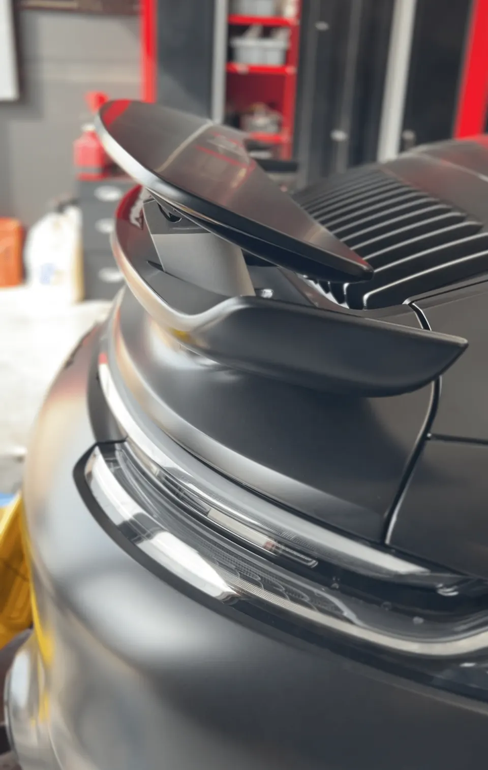 Close-up of the matte black rear of a Porsche, showcasing its active rear wing and LED taillight in a shop setting.