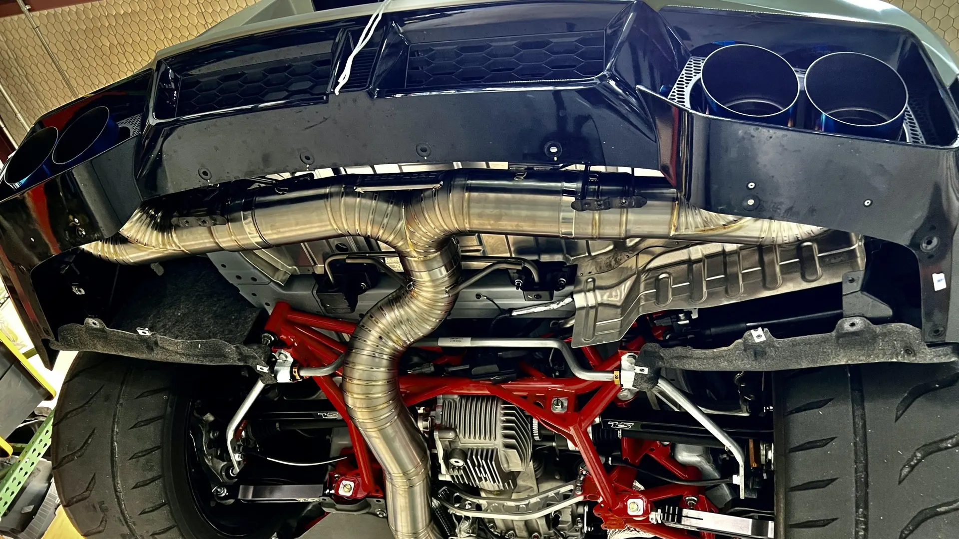 Underside view of a high-performance car on a lift, featuring a custom titanium exhaust system, rear differential, and red suspension.