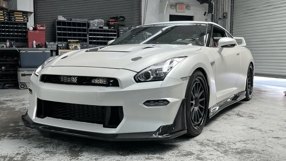 White Nissan GT-R with aftermarket front splitter, side skirts, and wheels parked inside a performance shop.