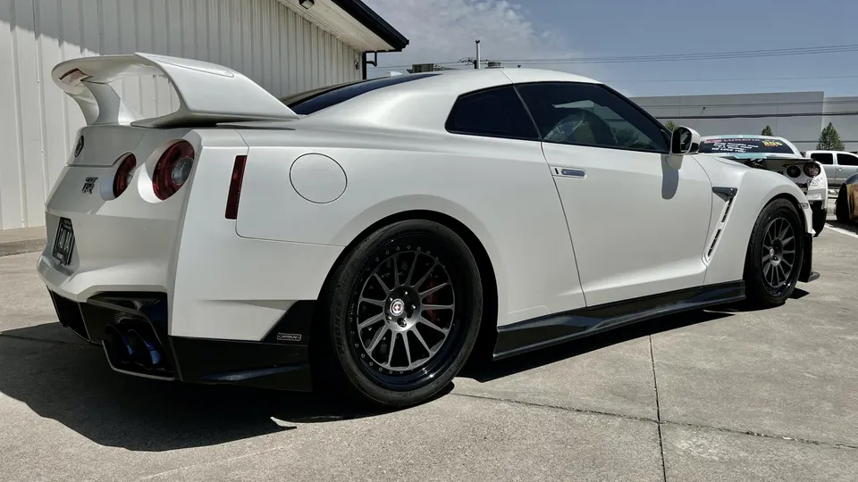 White Nissan GT-R with aftermarket wheels, large rear wing, and side skirts parked outdoors on a sunny day.