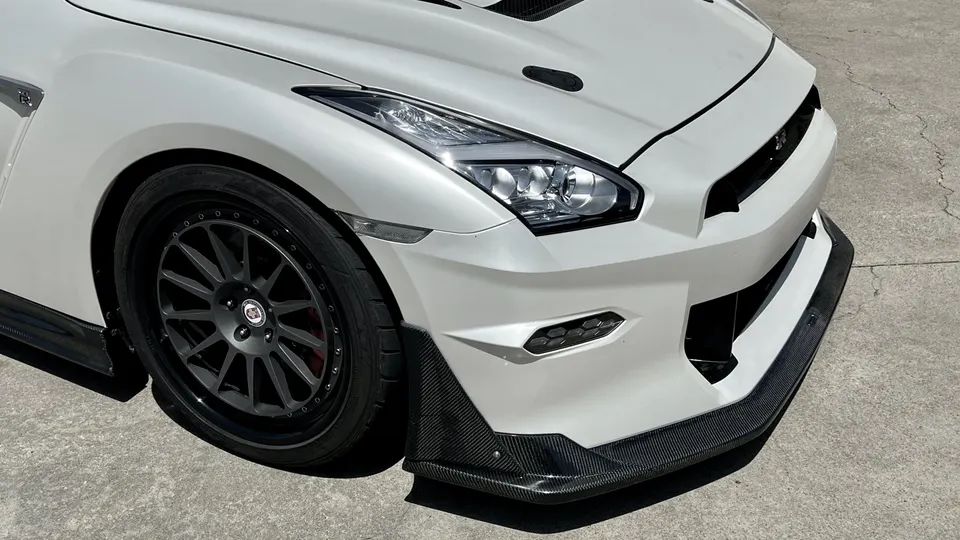 A white Nissan GT-R with aftermarket dark grey wheels and a carbon fiber front splitter.