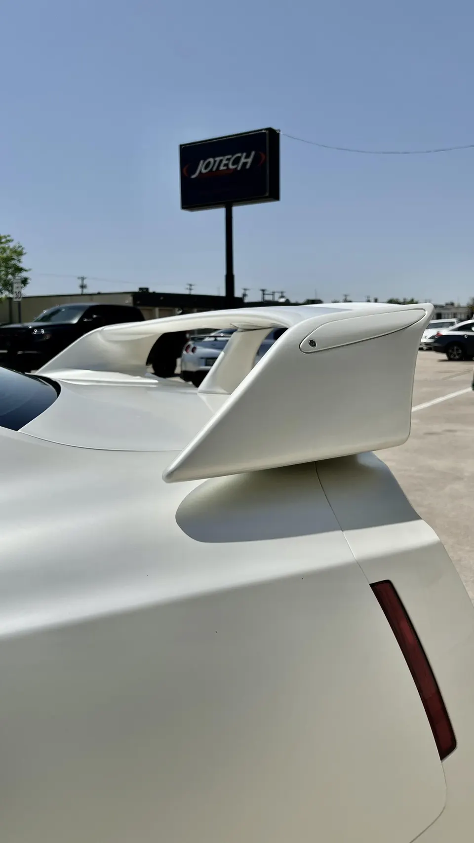 White sports car with a large aftermarket rear wing in a parking lot, with a Jotech sign visible in the background.