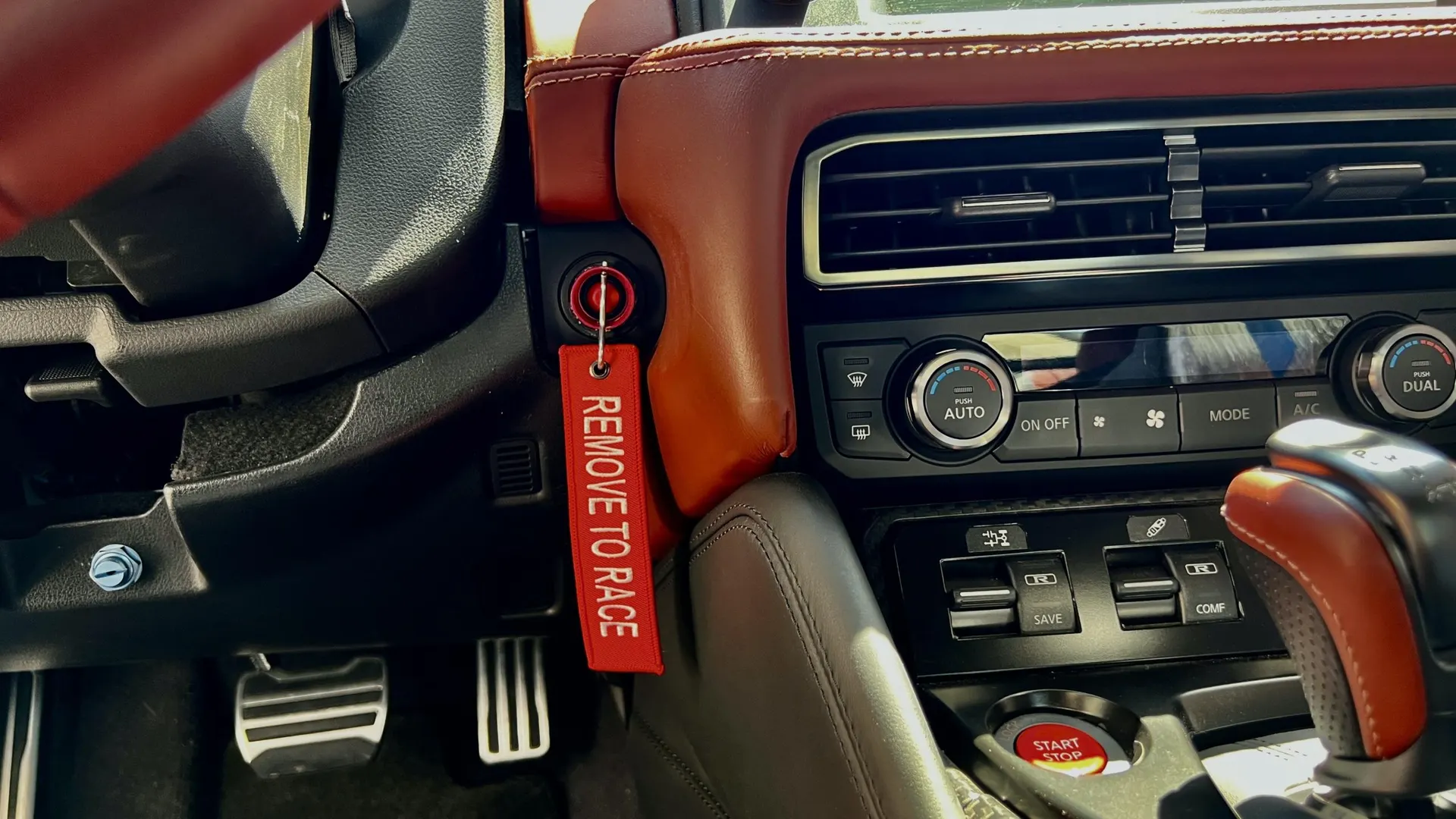 Close-up view of a car's interior, showing the dashboard, climate controls, pedals, shifter, and a red 'REMOVE TO RACE' keychain.