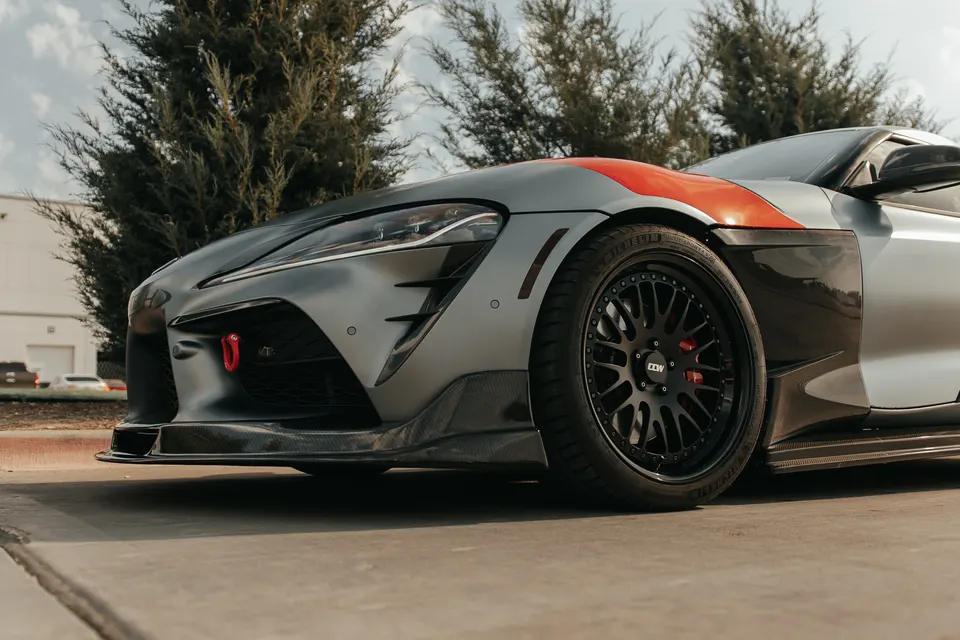 A matte grey Toyota Supra A90 with carbon fiber aero, custom black wheels, and a red tow hook parked outdoors.