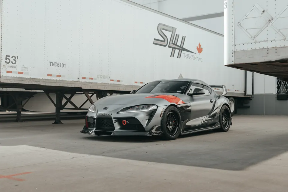 A grey Toyota Supra MK5 with an aftermarket aero kit and black wheels parked in an industrial setting.