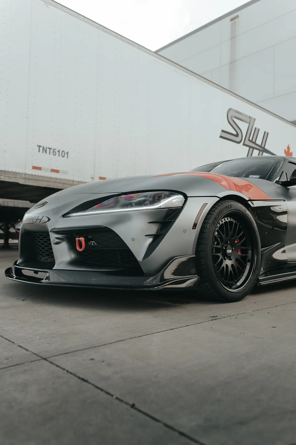 Modified Toyota Supra A90 with matte grey wrap, carbon fiber front splitter, custom black wheels, and red tow hook.