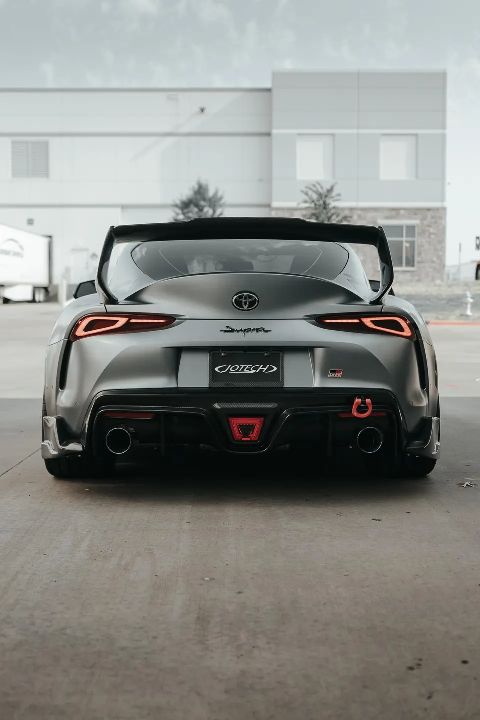 Rear view of a grey Toyota Supra MK5 with an aftermarket wing, diffuser, and exhaust tips, parked outside a building.