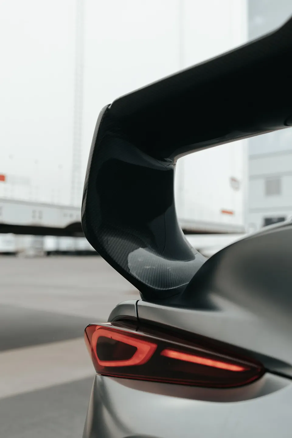 A close-up of a carbon fiber rear wing and LED taillight on a grey sports car.