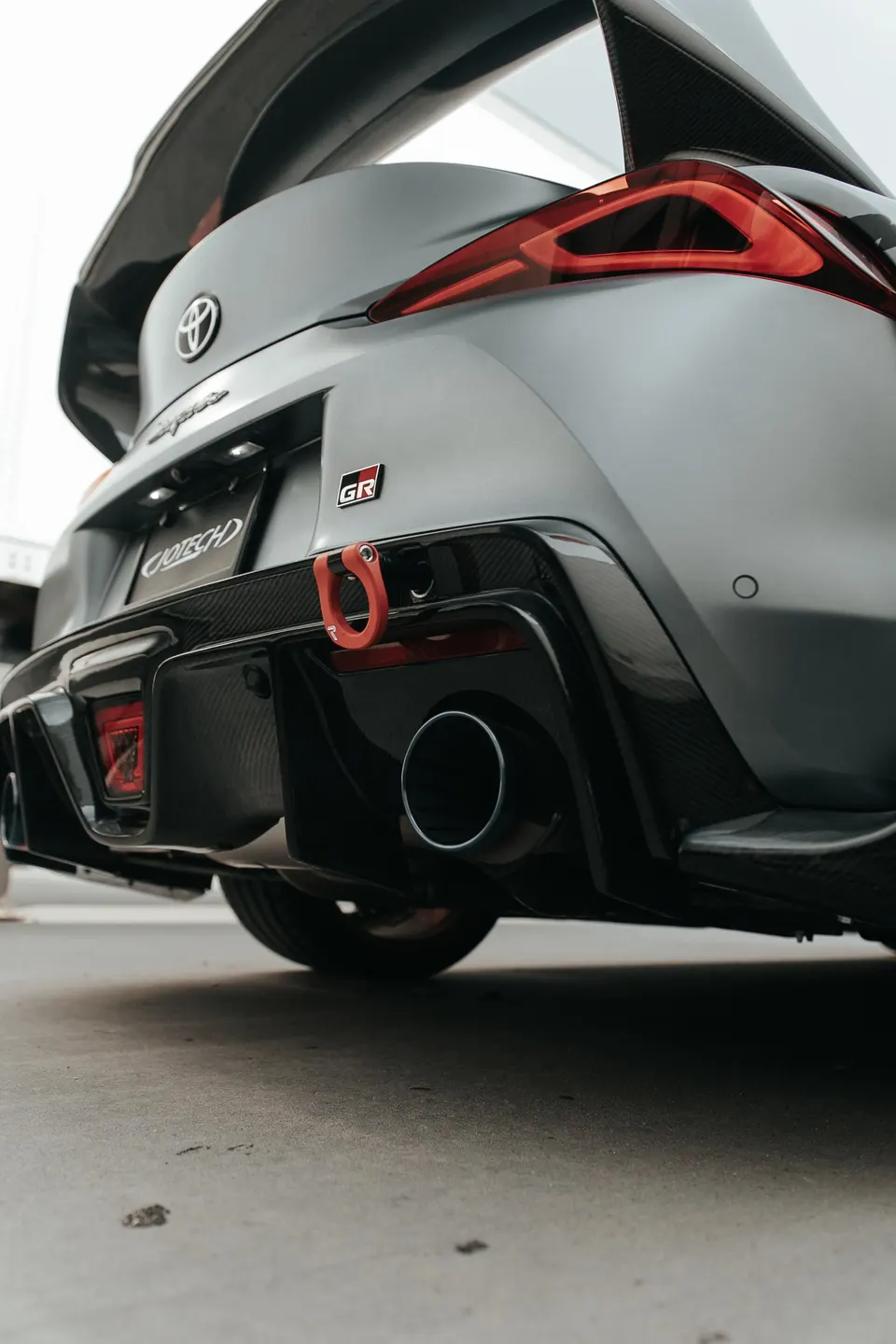 A grey Toyota GR Supra with a carbon fiber rear wing, diffuser, and a red tow hook, showcasing the exhaust tip.