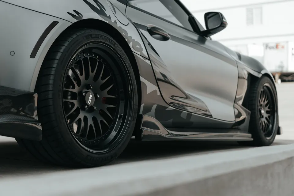 Grey Toyota Supra with widebody kit, side skirts, and black CCW wheels parked outdoors.