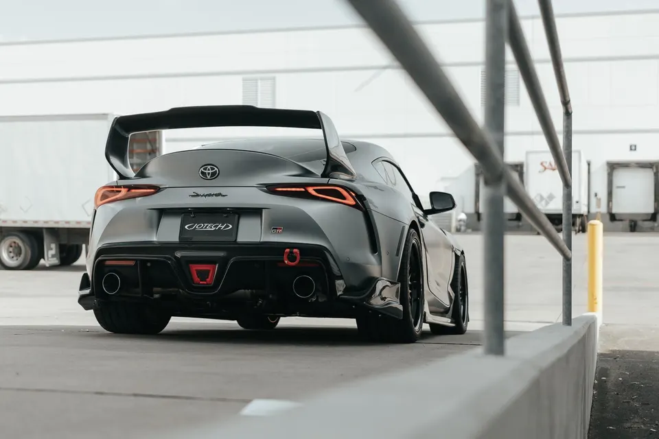 Rear view of a grey Toyota Supra MK5 with a large aftermarket wing, diffuser, and custom exhaust tips in an industrial setting.