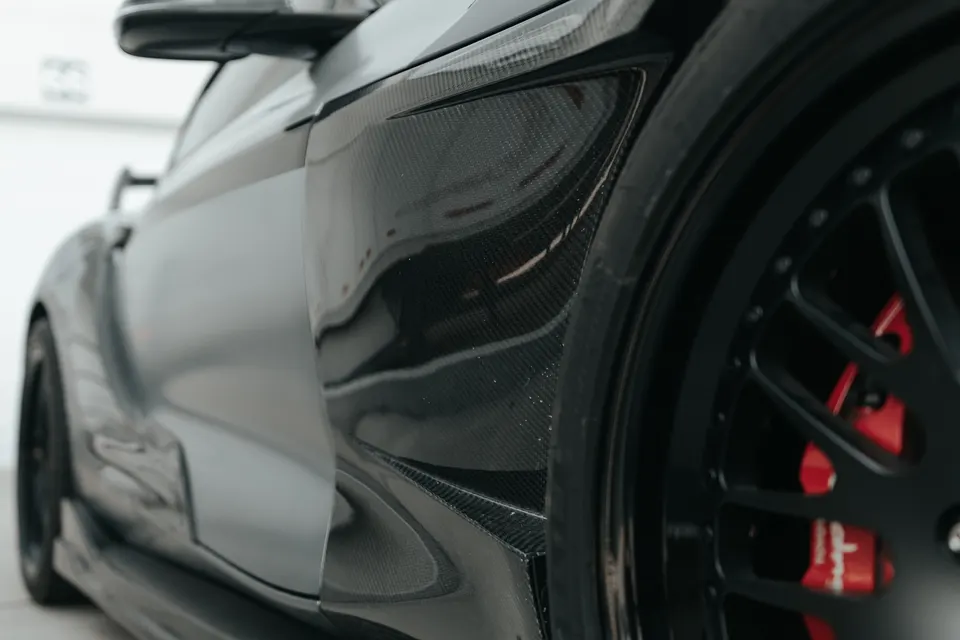 Close-up of a performance car's carbon fiber fender, black custom wheel, and red brake calipers.