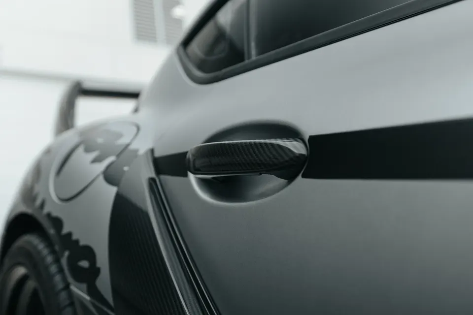 Close-up of a grey sports car's side, featuring a carbon fiber door handle and a subtle graphic on the rear fender.
