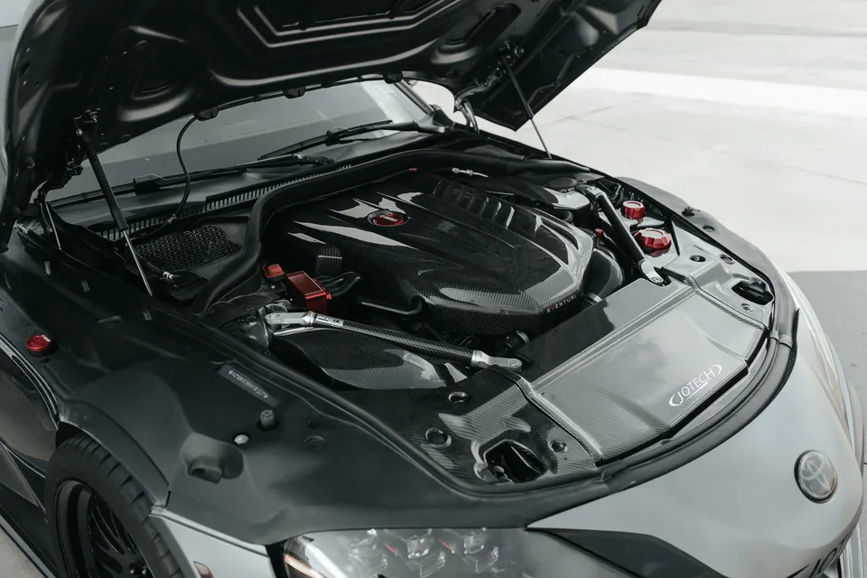 A close-up of a Toyota Supra engine bay featuring a carbon fiber Eventuri intake system and Jotech branding.