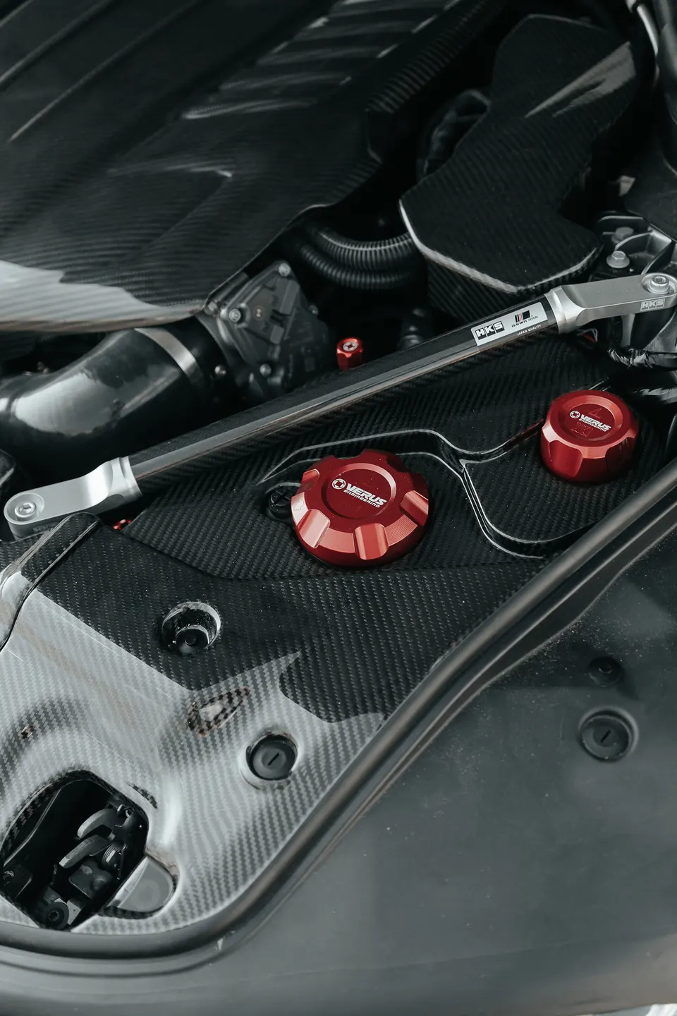 Close-up of a car's engine bay featuring carbon fiber components, an HKS strut bar, and red Verus Engineering caps.