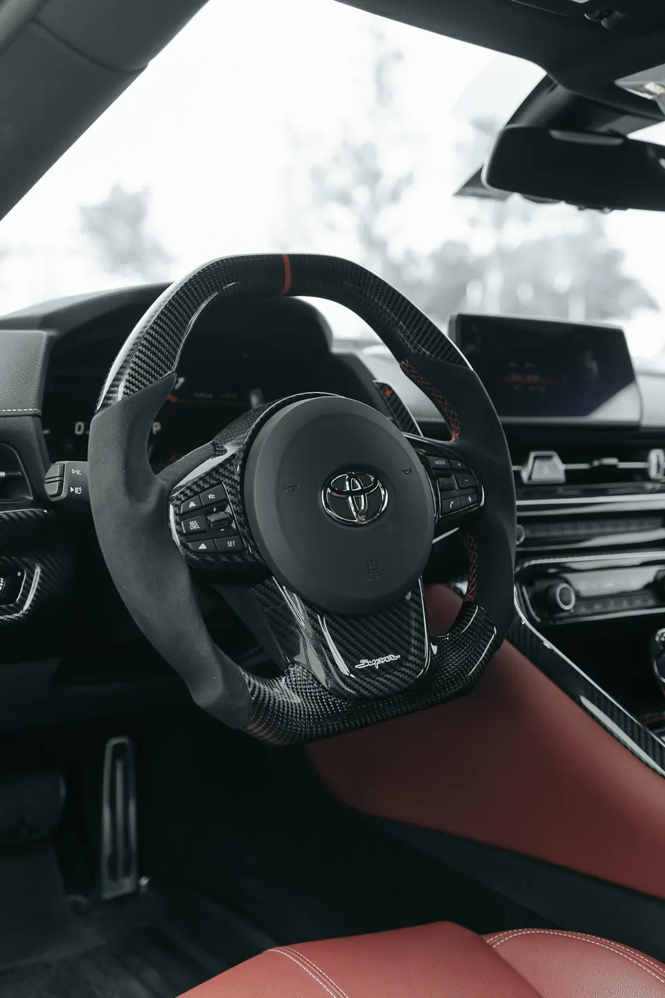 Close-up of a custom carbon fiber and Alcantara steering wheel with red stitching in a Toyota Supra interior.