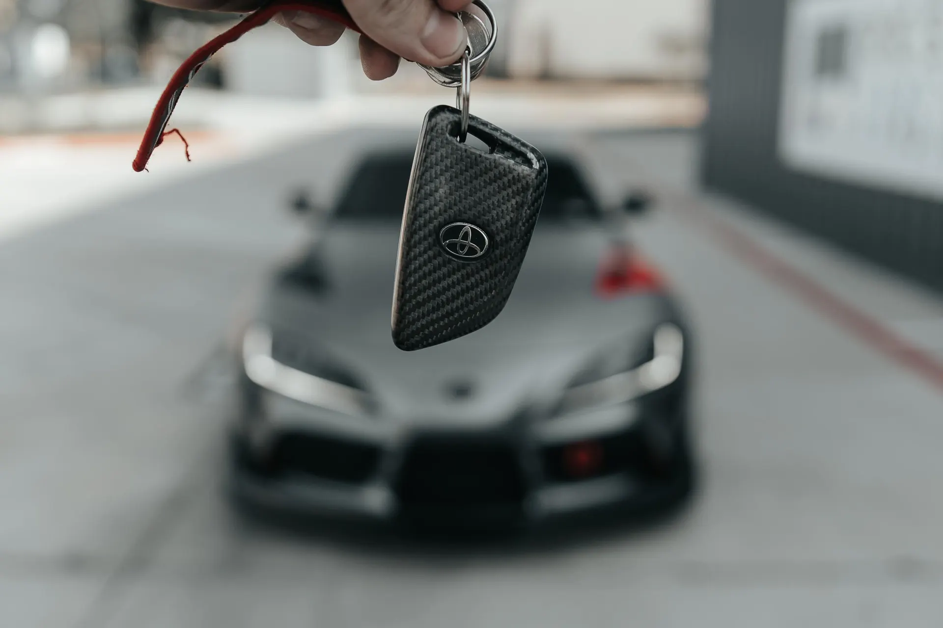 Hand holding a Toyota Supra car key with a carbon fiber cover, with the grey Supra blurred in the background.