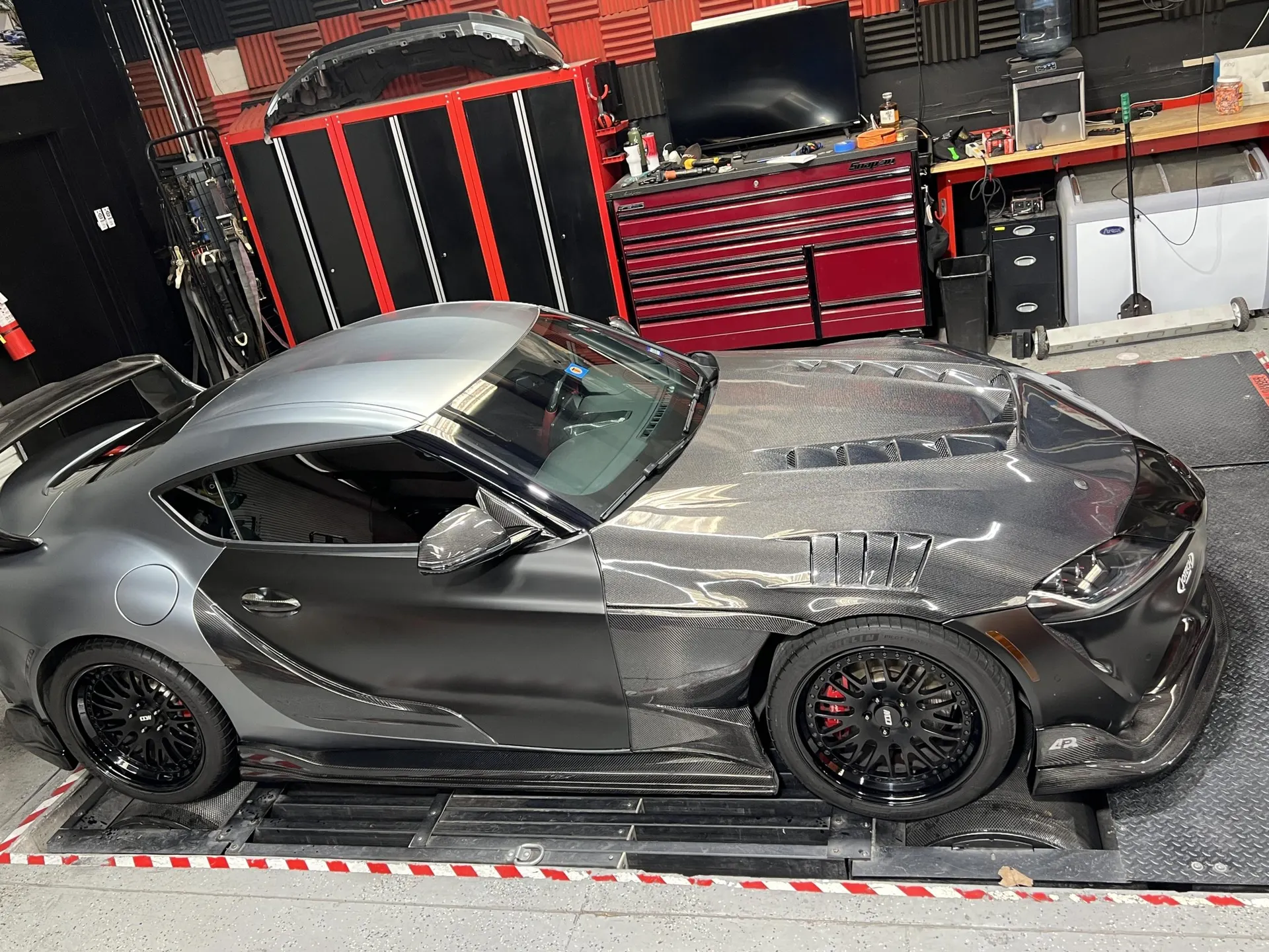 A custom widebody Toyota Supra on a dyno at a performance tuning shop, showcasing its carbon fiber bodywork and custom wheels.