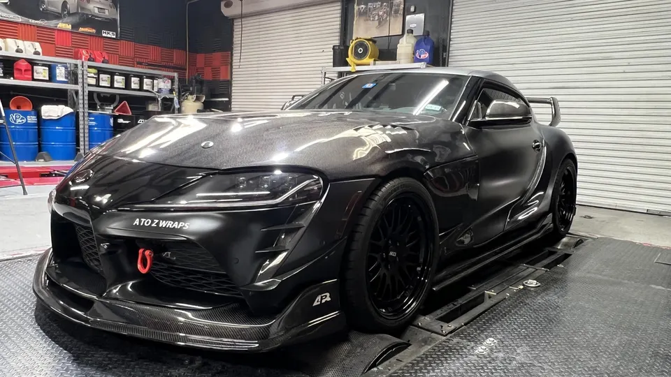 A black Toyota A90 Supra with carbon fiber body panels and a large rear wing on a dyno at a performance shop.