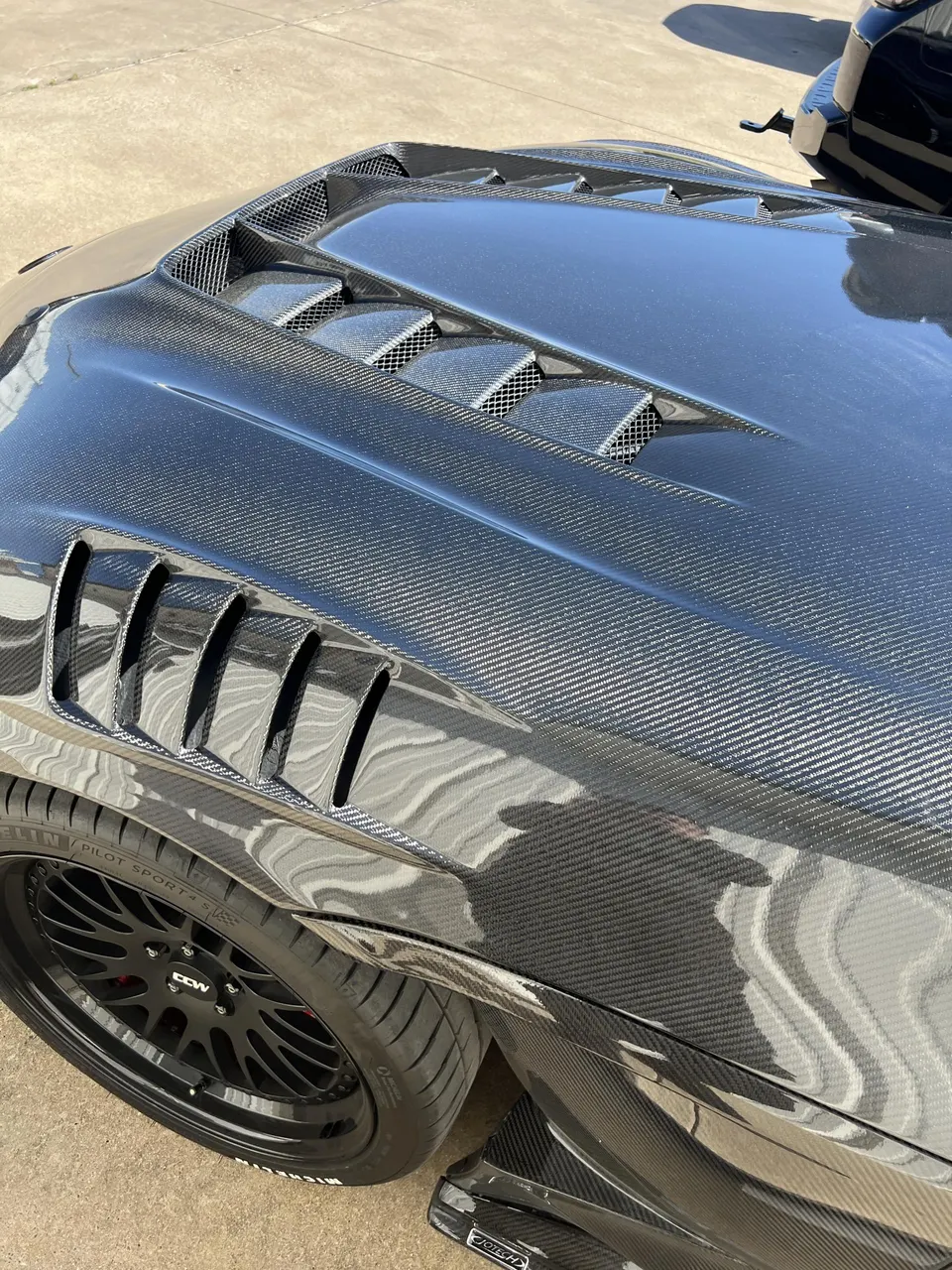 A close-up of a black carbon fiber car hood and fender with vents, showcasing CCW wheels and Michelin tires.