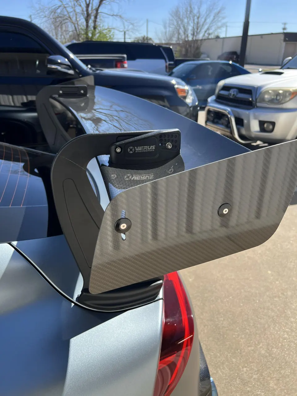 Close-up of a carbon fiber rear wing with Verus Engineering branding on a grey car, showcasing aerodynamic details.