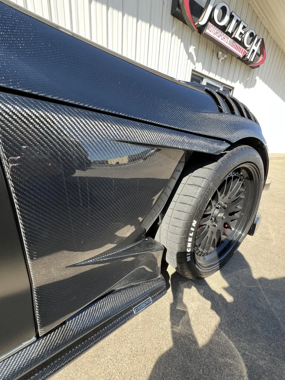 Close-up of a black carbon fiber widebody car with custom wheels and Michelin tires outside Jotech Motorsports.