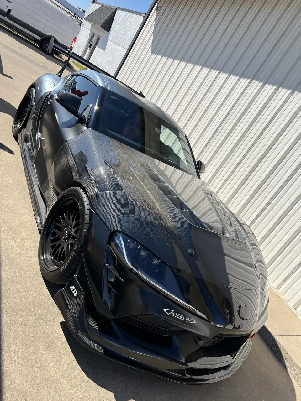 Highly modified gray Toyota A90 Supra with carbon fiber widebody kit, aftermarket wheels, and custom aero parked outdoors.
