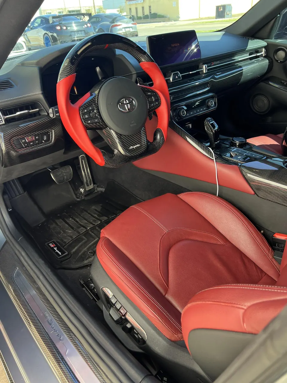 Interior view of a Toyota Supra featuring a custom carbon fiber and red leather steering wheel, red leather seats, and dashboard.
