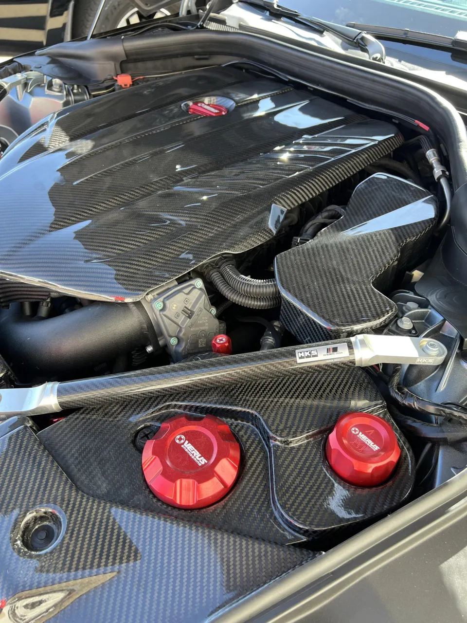 Close-up of a performance car engine bay featuring a carbon fiber engine cover, HKS strut bar, and Verus Engineering fluid caps.