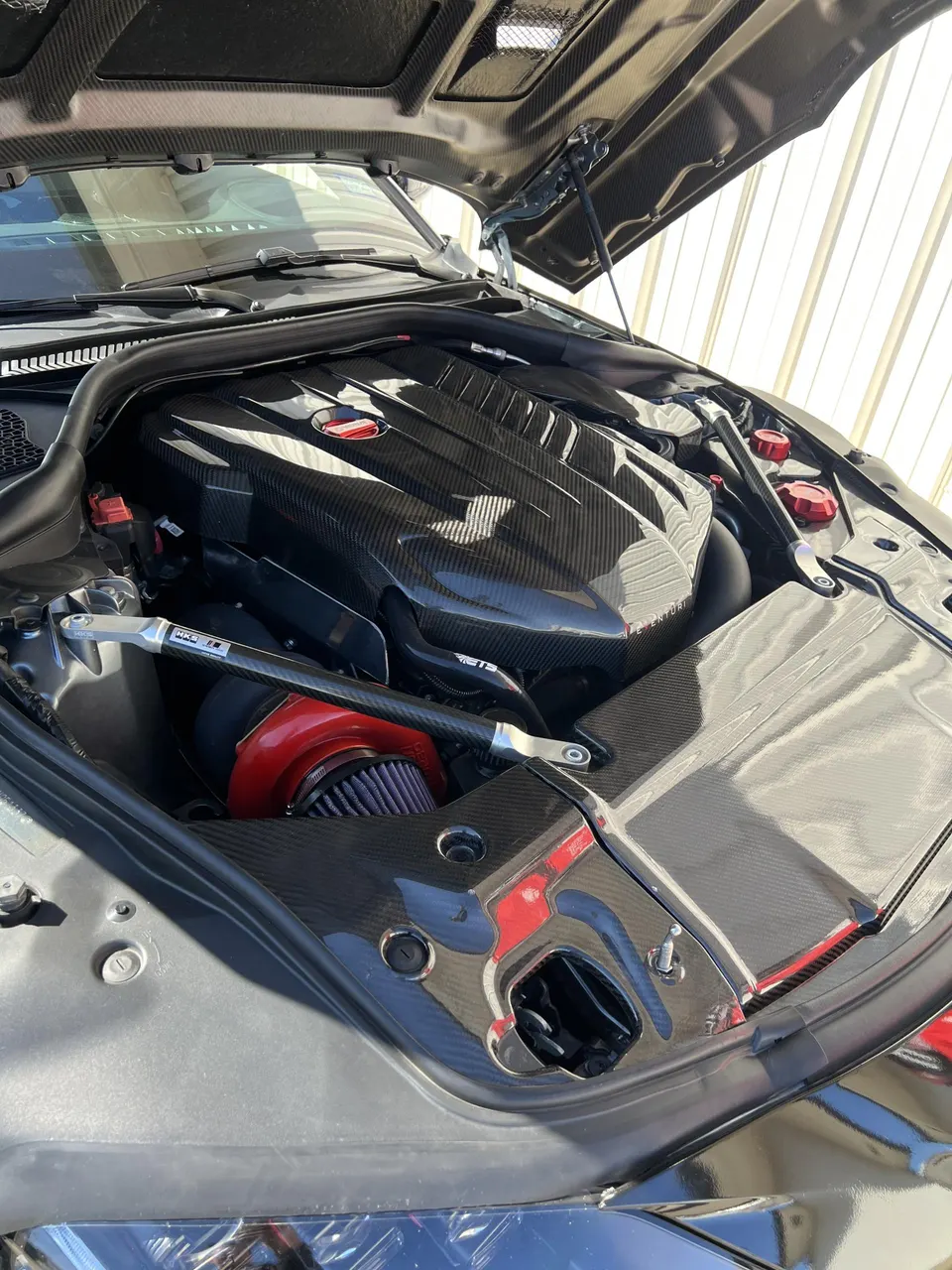 Close-up of a modified Toyota Supra engine bay with carbon fiber engine cover, aftermarket intake, and strut bar.