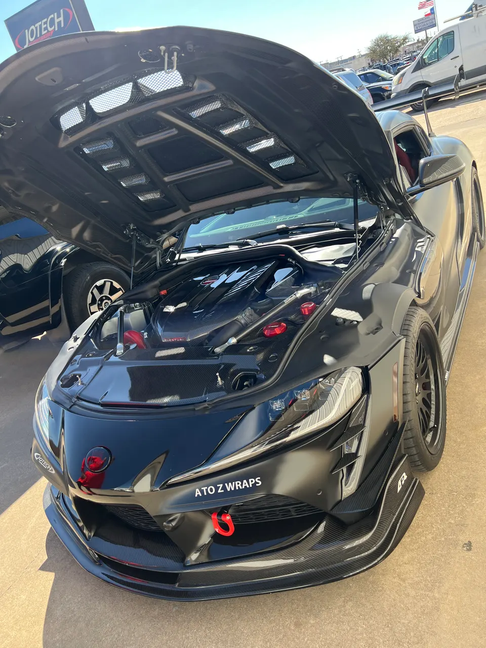 Modified black Toyota Supra with carbon fiber vented hood open, showcasing engine bay and front aero with red tow hook.
