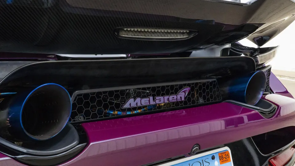 A close-up of the rear of a purple McLaren, showcasing blue-tipped exhaust, carbon fiber diffuser, and rear wing.