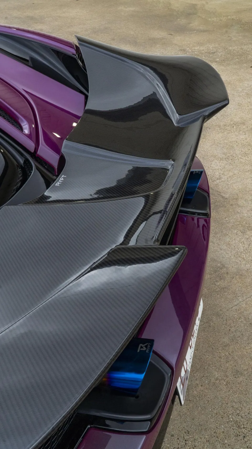 Close-up of a purple McLaren's rear, showcasing a glossy carbon fiber rear wing and blue-tipped exhaust outlets.