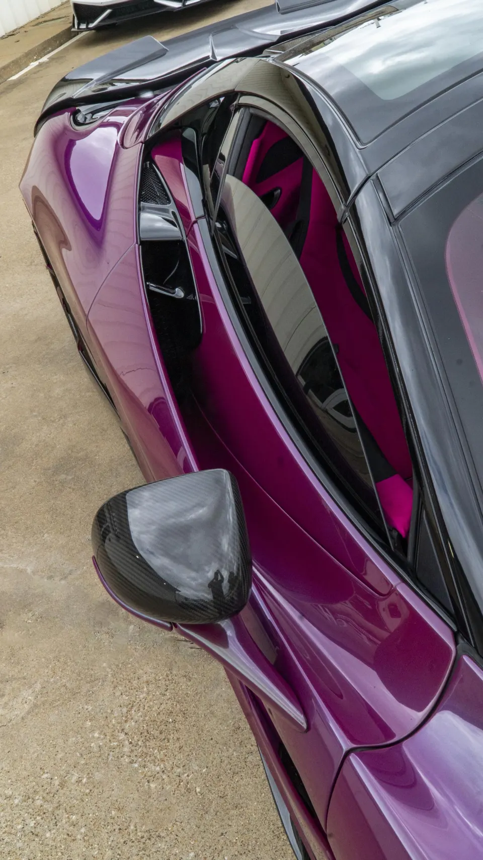 A high-angle view of a vibrant purple McLaren sports car, featuring a carbon fiber side mirror and a bright magenta interior.