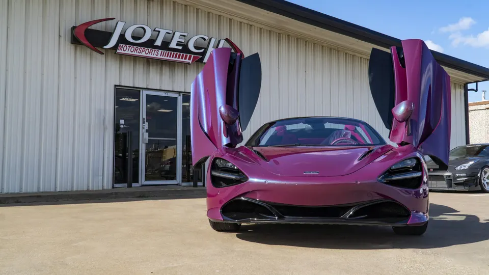 A vibrant purple McLaren 720S with its butterfly doors open, parked prominently in front of the Jotech Motorsports building.