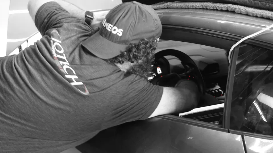 A Jotech mechanic wearing an Eneos hat works on the interior electronics of a performance car, interacting with the dashboard.