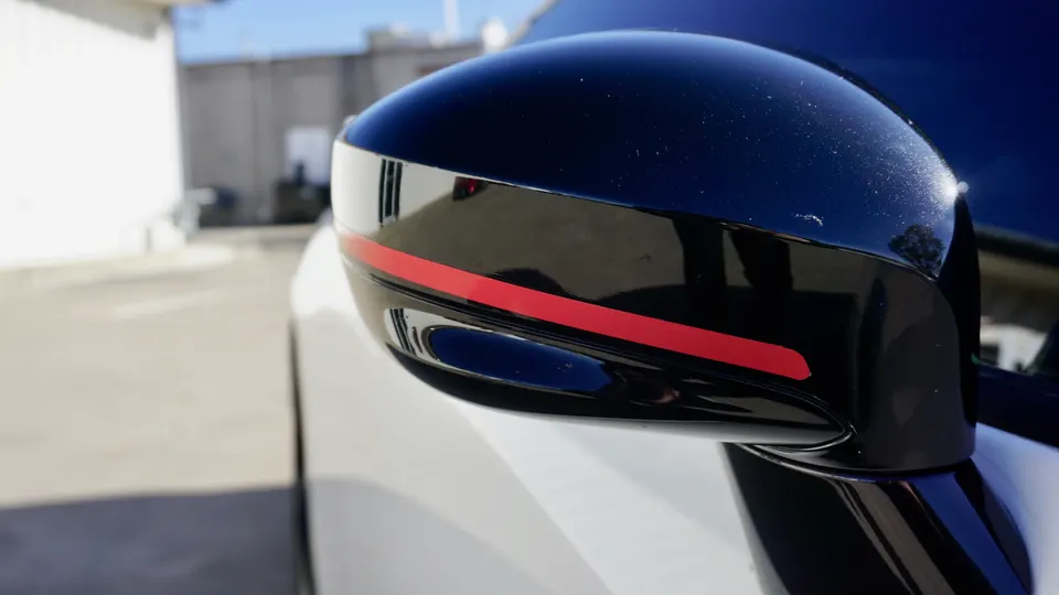 Close-up of a black car side mirror with a red accent stripe, showcasing exterior detail on a sunny day.
