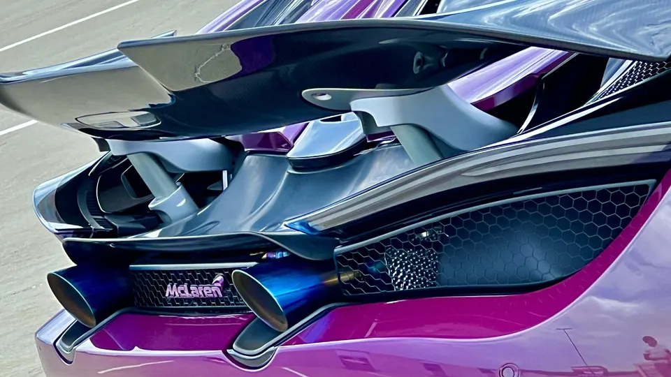 A close-up of the rear of a purple McLaren, showcasing its carbon fiber rear wing, diffuser, and blue exhaust tips.