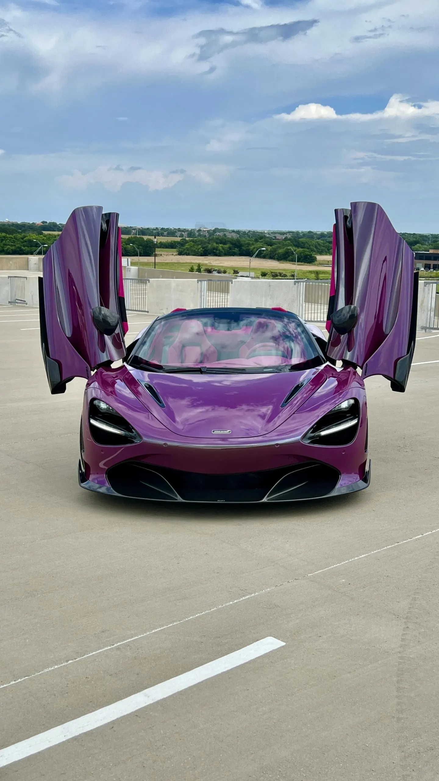 A vibrant purple McLaren 720S with its butterfly doors open, parked on a concrete lot under a cloudy sky.