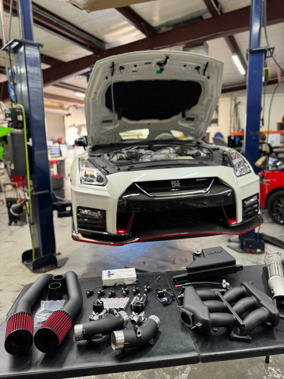 White Nissan GT-R on a lift in a performance shop with aftermarket induction, turbo, and fuel system parts laid out.