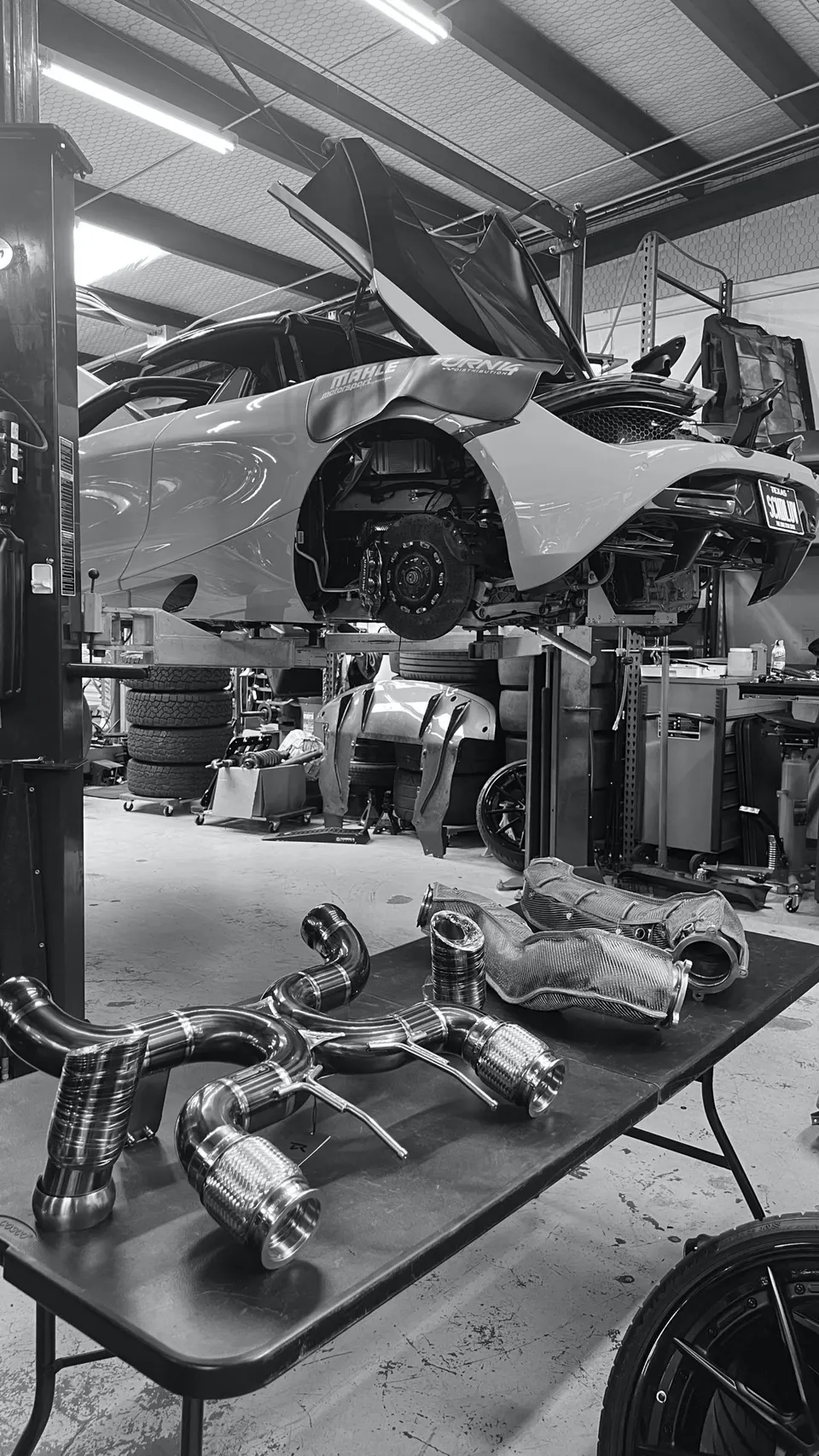 A McLaren supercar on a lift in a performance shop, with a disassembled exhaust system and carbon fiber components on a table.
