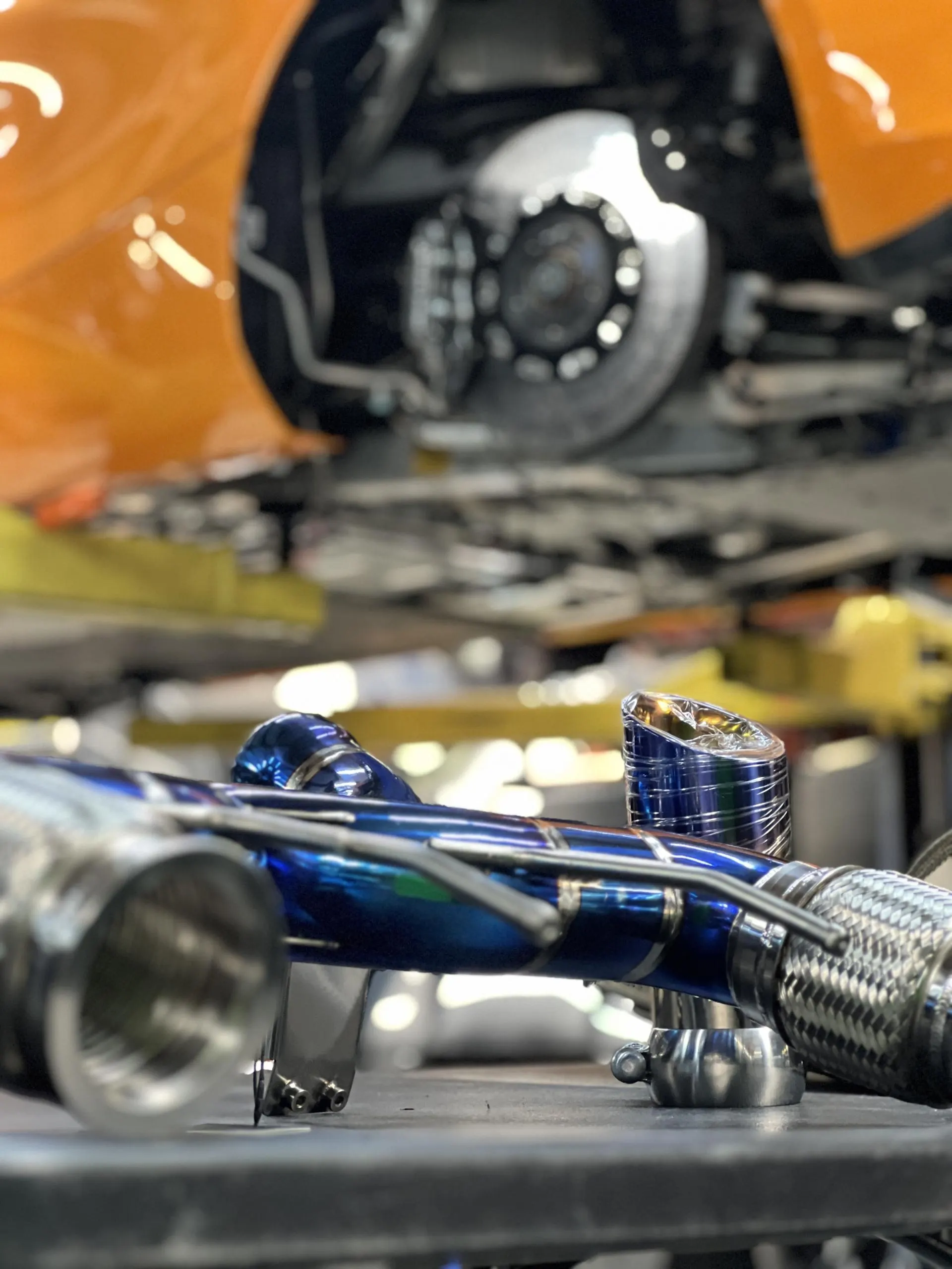 Titanium exhaust component on a workbench in a performance tuning shop with an orange car on a lift in the background.