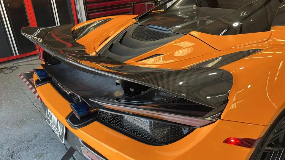 Rear view of an orange McLaren 720S with a carbon fiber rear wing and blue-tipped exhaust in a performance shop.