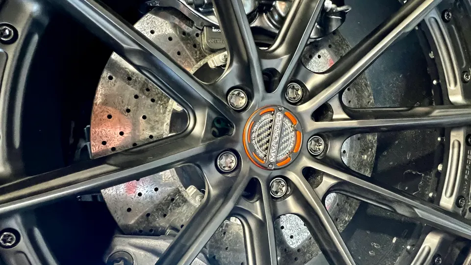 A close-up of a dark gray MV Forged wheel with a carbon fiber center cap and drilled brake rotors.