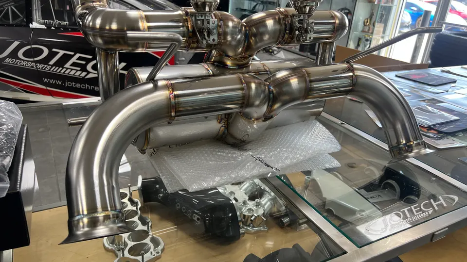 A high-performance titanium exhaust system with intricate welds on display at Jotech Motorsports, with a race car in the background.