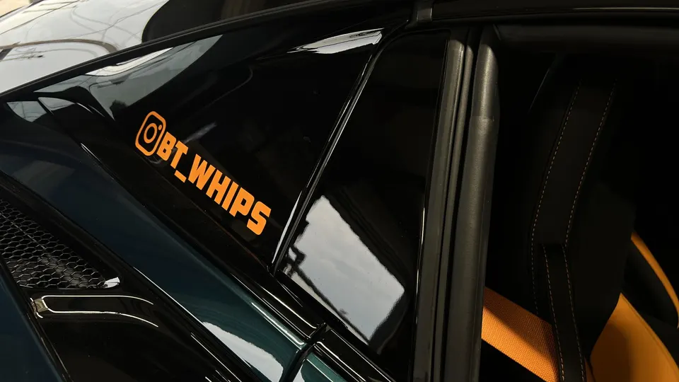 A close-up of a black car's rear side window with an orange Instagram sticker and a glimpse of the orange interior.