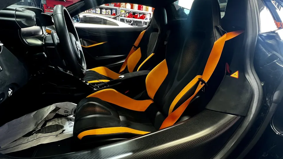Interior view of a McLaren with black and orange racing seats, orange harnesses, carbon fiber trim, and steering wheel.