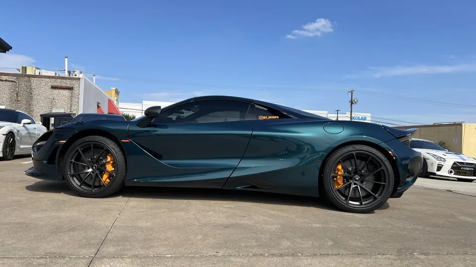 Dark green McLaren 720S supercar with black wheels and orange brake calipers parked outdoors, with a white Nissan GT-R in the background.
